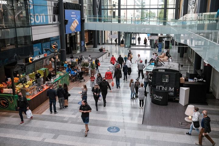 Ett köpcenters inomhusområde där flera människor rör sig. Till vänster finns ett caféområde och till höger en liten försäljningsstånd