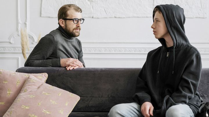 En illustration. En vuxen person samtalar med en ungdom som bär en hoodie i en soffa.