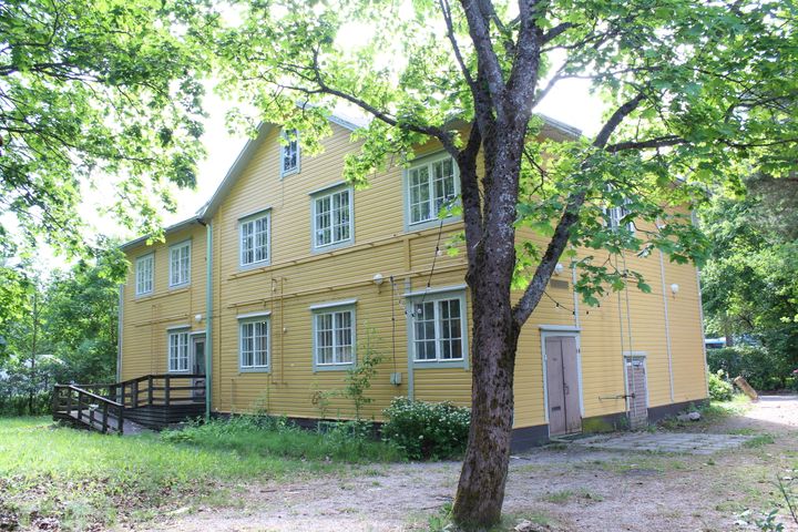 Kaksikerroksinen keltainen puutalo vehreässä ympäristössä.