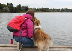 Tyttö istuu laiturilla järven rannalla pinkki takki yllään, selässään punainen reppu. Hän on kumartuneena ruskeavalkoisen koiran puoleen.