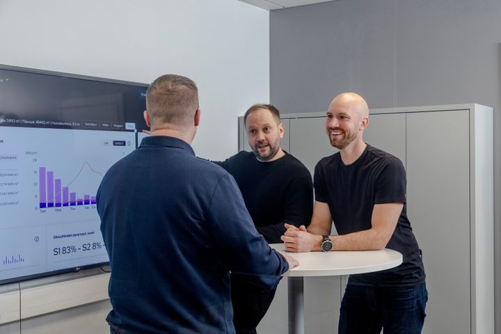 Kolme henkilöä keskustelemassa toimistossa korkean pöydän ääressä. Yksi heistä osoittaa seinänäyttöä, jossa on kaavioita.