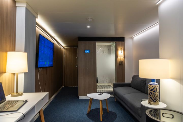 Alt-teksti: Moderni laivan hytti, jossa on sohva, pieni pöytä, televisio ja lamppuja.