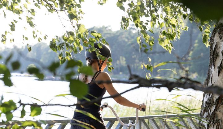 Henkilö polkee pyörällä vehreässä puistossa, taustalla näkyy vettä ja puita.