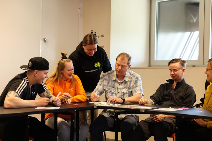 Iloiset ihmiset keskustelevat koulutustilaisuudessa pöydän ääressä.