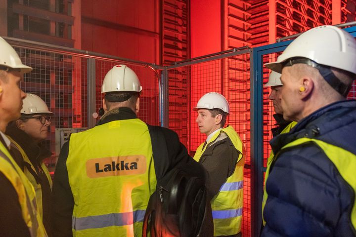 Pieni ryhmä ihmisiä keltaisissa huomioliiveissä ja kypärissä, seisomassa teollisuusympäristössä.