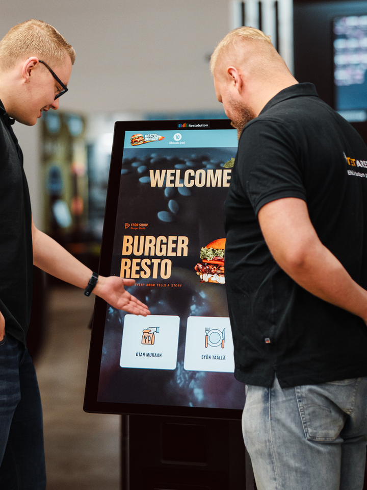 Kaksi henkilöä katsoo ravintolan itsepalvelukassaa, jossa on tervetuloa-viesti ja valikko.