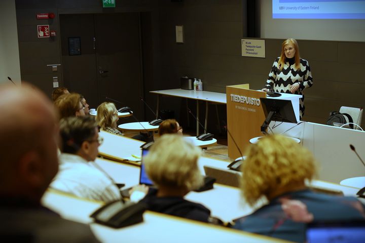 Nainen puhuu yleisön edessä salissa.