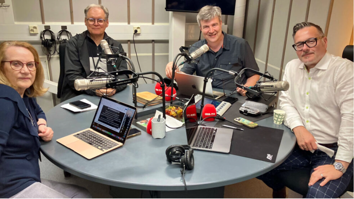 Ryhmä ihmisiä istuu pöydän ääressä mikrofonien kanssa radionauhoitushuoneessa.