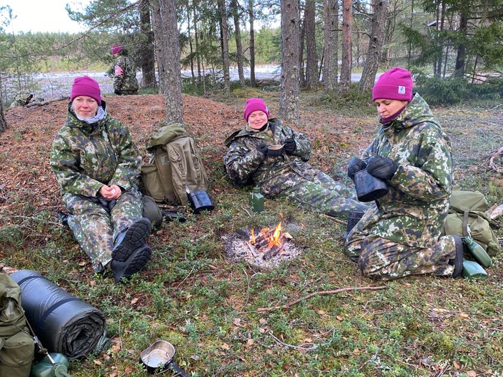 Kolme naista maastopuvuissa istumassa nuotion äärellä metsässä.