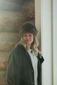 A woman wearing a furry hat and coat stands indoors by a wooden wall.
