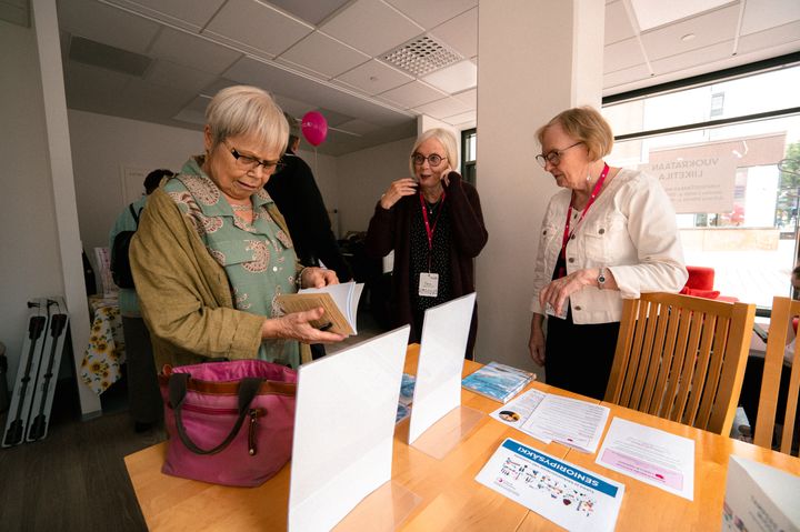Kolme naista tutustuu Senioripäivän esitteisiin pöydän ääressä TampereMission talolla.
