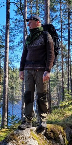 Kuvassa Ali Leiniö metsässä, reppu selässä, takana puiden välistä siintää sininen taivas